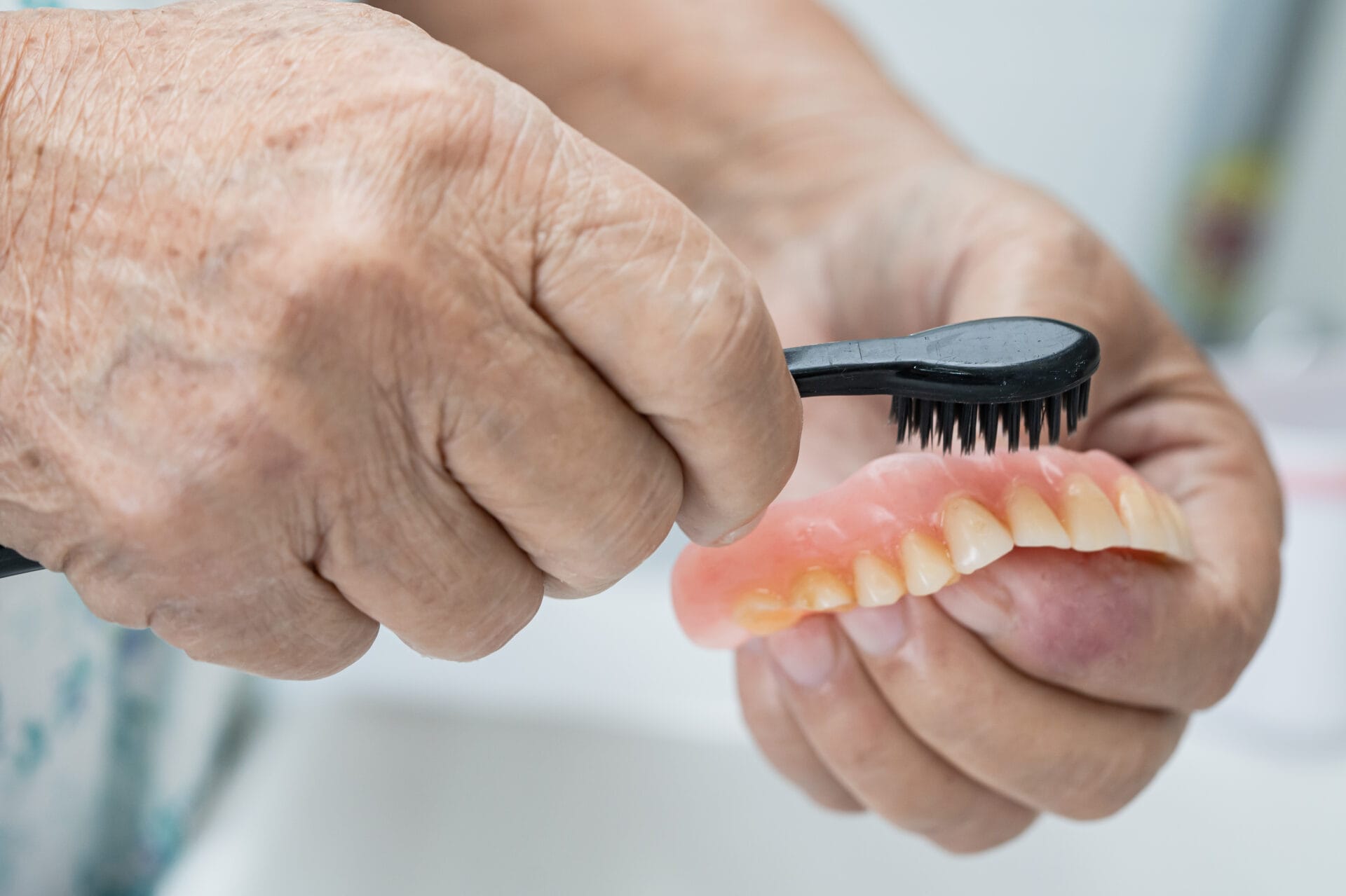 Asian senior woman patient using a toothbrush to clean dentures, demonstrating proper care and maintenance.